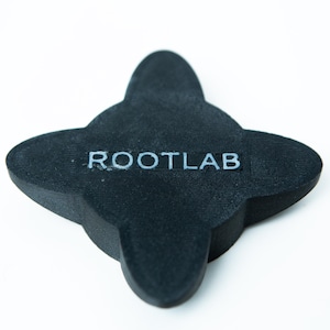 May include: A black, four-lobed object with the text "ROOTLAB" in white. The object has a matte finish and is set against a white background. The shape resembles a stylized flower or emblem.