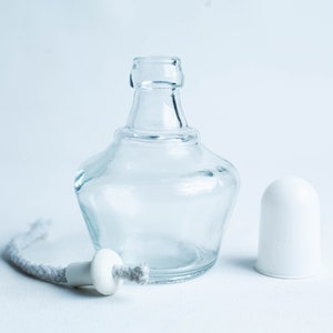 May include: A clear glass bottle with a white ceramic stopper and a white wick. The bottle is empty and has a round shape.