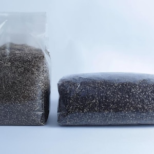 May include: Two clear plastic bags filled with dark, granular material. One bag is upright and rectangular, while the other is lying horizontally. The contents appear to be a type of seed or grain, with a speckled appearance.