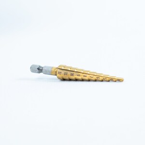 May include: A gold-coloured step drill bit with a silver hex shank. The bit has numbers 4 through 12 printed on the side.