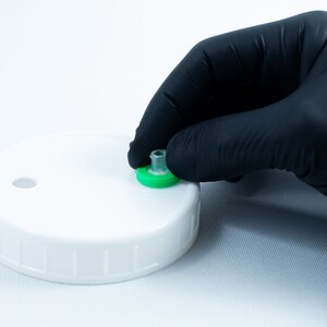 May include: A white plastic lid with a green ring and a small clear tube. A gloved hand is holding the tube.