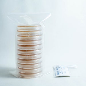 May include: A stack of clear, round petri dishes with a light brown agar inside, sealed in a clear plastic bag. A small packet of desiccant is next to the bag. The background is plain white.