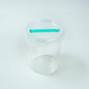 May include: A clear, cylindrical plastic container with a matching transparent lid. The lid features a rectangular slot across the centre, which is accented by a bright teal, serrated-edge plastic insert for dispensing items through the top.