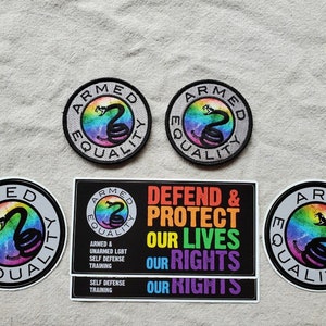 Könnte beinhalten: Drei Armed Equality Aufkleber mit einem Regenbogen-Schlangen-Design. Ein Aufkleber ist rund mit dem Text "Armed Equality" in einem schwarzen Kreis. Die beiden anderen Aufkleber sind rechteckig mit dem Text "Armed & Unarmed LGBT Self Defense Training" und "Defend & Protect Our Lives Our Rights Our Rights".