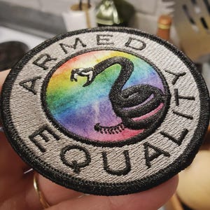 May include: Circular embroidered patch with the words "ARMED EQUALITY" in a silver border. The centre shows a black swan against a rainbow gradient background.