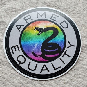 Puede incluir: Una pegatina redonda con un borde negro y un fondo gris. La pegatina dice "ARMED EQUALITY" en un círculo negro alrededor de un fondo de colores arcoíris con una serpiente de cascabel negra enroscada en la parte superior.