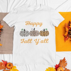 May include: White t-shirt with the text "Happy Fall Y'all" in orange lettering. Three orange pumpkins with a leopard print design are above the text.