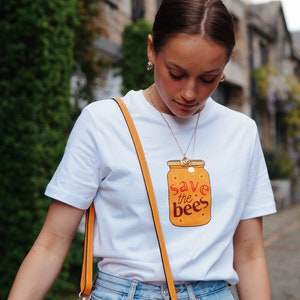 May include: White t-shirt featuring a cartoon illustration of a jar of honey with the text "save the bees" in orange and red. The shirt has short sleeves and is being worn with a yellow shoulder bag.