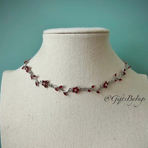 May include: A silver necklace with a delicate vine design featuring small red flowers. The necklace is displayed on a white mannequin bust.