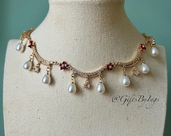 Puede incluir: Un collar dorado con un diseño delicado, con pequeños acentos florales rojos, cristales transparentes y perlas colgantes en forma de lágrima. El collar se exhibe en un maniquí. El texto "@GiftsByIngs" es visible.