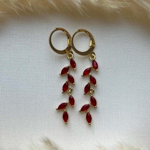 May include: A pair of gold hoop earrings with dangling red gemstone drops. The earrings have a simple design and are perfect for everyday wear.