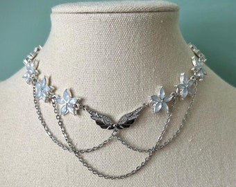 May include: A silver necklace with a delicate chain and multiple layers. The necklace features small white flower-shaped charms and a central butterfly design.