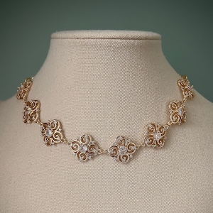 Peut inclure: Un collier en or avec un design délicat et orné, orné de petites pierres claires. Le collier est présenté sur un mannequin recouvert de tissu blanc.