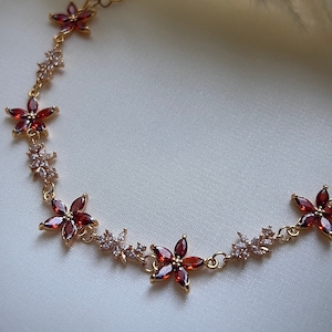 Puede incluir: Un collar de cadena dorada con acentos de flores de cristal rojo y transparente.