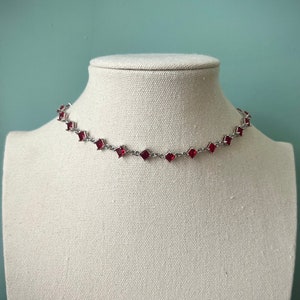 May include: A silver chain choker necklace with red square-shaped rhinestones.