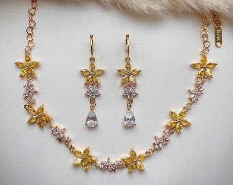 May include: A gold necklace and earrings set with yellow and clear cubic zirconia stones. The necklace features a delicate chain with flower-shaped pendants. The earrings are drop earrings with a flower-shaped design.