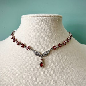 May include: Silver necklace with a red gemstone pendant shaped like wings. The necklace is adorned with small red flower-shaped accents.