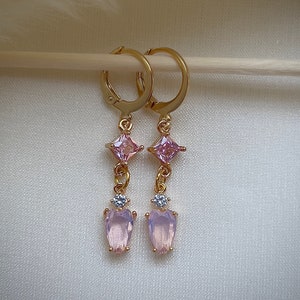 Puede incluir: Aretes de aro dorados con un colgante de cristal rosa y transparente. El colgante tiene un cristal cuadrado, un cristal redondo más pequeño y un cristal en forma de corazón.