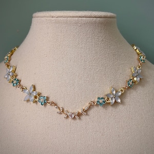 May include: A gold-coloured necklace with a floral design. It features light blue and white flower accents, small turquoise flowers, and clear crystals. The necklace is displayed on a mannequin.