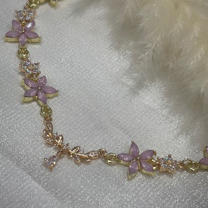 May include: A gold chain bracelet with pink flower charms and clear crystal accents. The bracelet is delicate and feminine.
