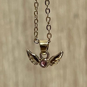May include: A gold chain necklace with a small pendant featuring a pair of angel wings with a pink gemstone in the center.