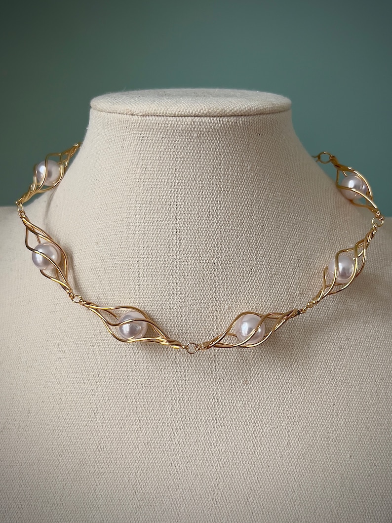May include: A gold wire necklace with a series of white pearls set in the wire. The necklace is on a white mannequin.