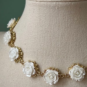 May include: A gold chain necklace with white rose-shaped charms. The necklace is on a white fabric mannequin.