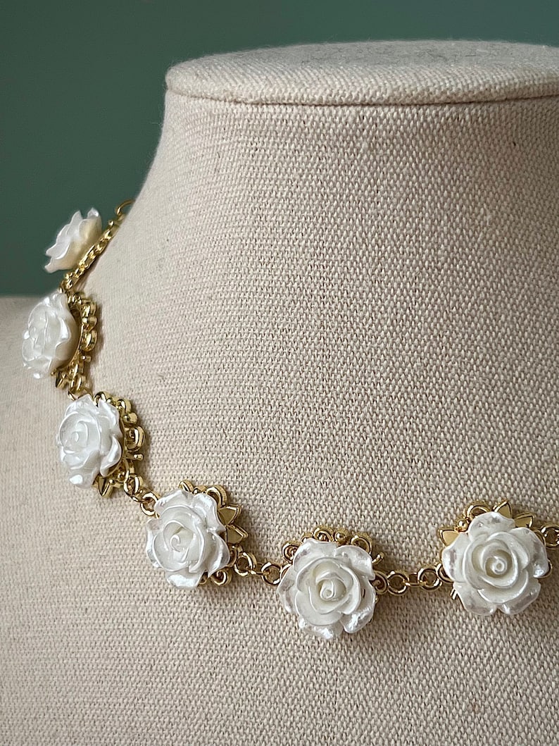 May include: A gold chain necklace with white rose-shaped charms. The necklace is on a white fabric mannequin.