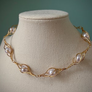 May include: A gold necklace with a delicate wire design that holds white pearls. The necklace is displayed on a white fabric mannequin.