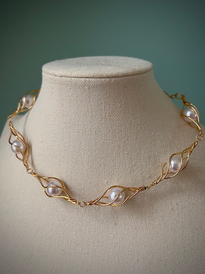 May include: A gold necklace with a delicate wire design that holds white pearls. The necklace is displayed on a white fabric mannequin.