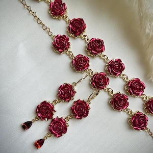 Op de afbeelding: Een set roosvormige sieraden met een paar oorbellen en een armband. De oorbellen hebben drie roosvormige bedels met rode edelsteenaccenten. De armband heeft een gouden ketting met meerdere roosvormige bedels.