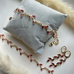 Könnte beinhalten: Ein Gold-Set aus Halskette, Armband und Ohrringen mit einem zarten Rankendesign und roten Edelstein-Akzenten. Der Schmuck ist auf einem grauen Samtkissen präsentiert. Der Text "@GiftsByIngs" ist im Bild sichtbar.