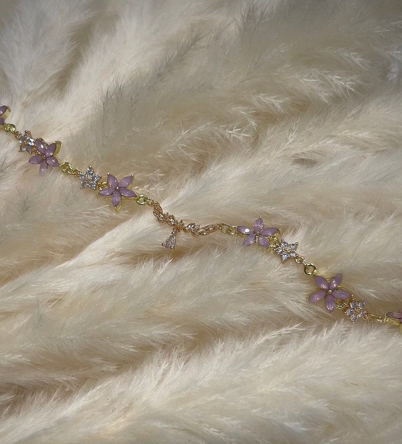 May include: A delicate gold chain bracelet with small pink flower charms and clear crystal accents. The bracelet is on a white fluffy background.