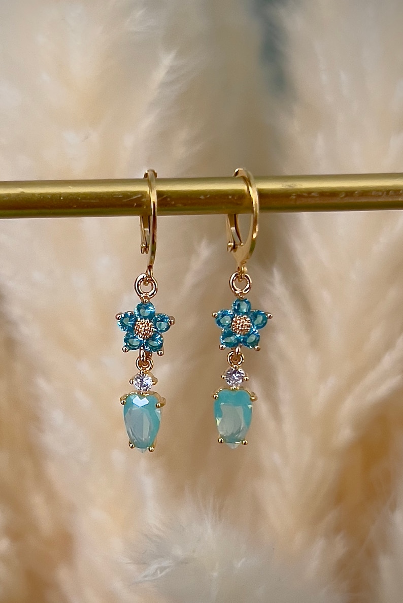 Puede incluir: Pendientes de aro dorados con un dije de flor azul claro y una piedra azul claro en forma de l&aacute;grima.