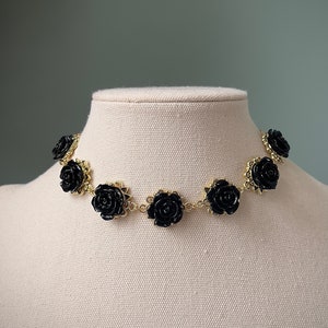 May include: A gold chain choker necklace with black rose-shaped charms. The necklace is displayed on a white mannequin.