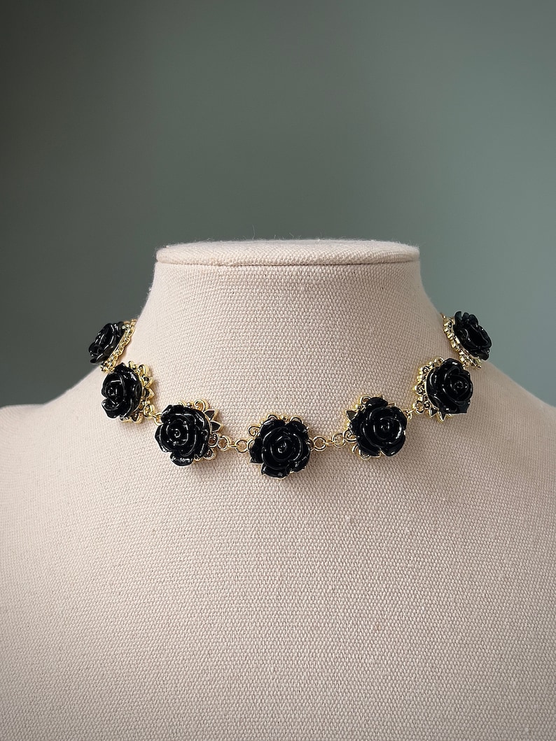May include: A gold chain choker necklace with black rose-shaped charms. The necklace is displayed on a white mannequin.