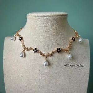 May include: A gold-toned necklace with a floral design, black accents, and dangling white teardrop-shaped pearls. The necklace is displayed on a mannequin. The necklace has the text "@GiftsByIngs".