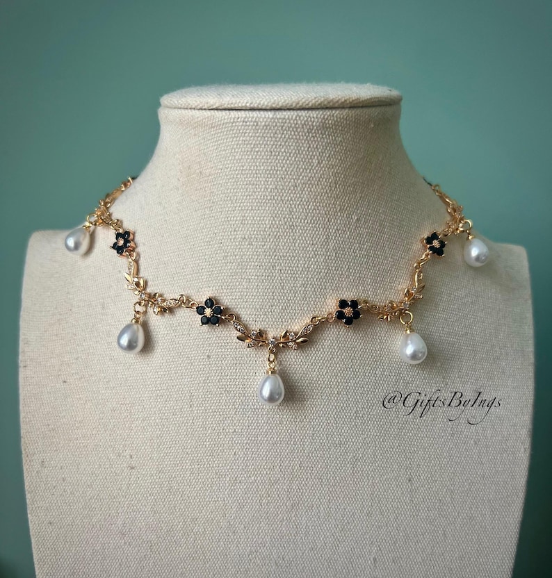 May include: A gold-toned necklace with a floral design, black accents, and dangling white teardrop-shaped pearls. The necklace is displayed on a mannequin. The necklace has the text "@GiftsByIngs".