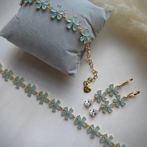 May include: A set of delicate gold jewelry featuring light blue flower-shaped charms. The set includes a bracelet, necklace, and earrings with dangling clear crystal drops.