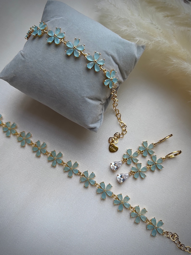 May include: A set of delicate gold jewelry featuring light blue flower-shaped charms. The set includes a bracelet, necklace, and earrings with dangling clear crystal drops.