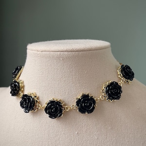 May include: A gold chain necklace with black rose-shaped charms. The necklace is on a white mannequin bust.