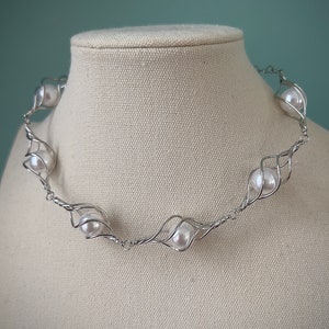 May include: A silver wire necklace with white pearls. The necklace is made of twisted wire that forms a delicate, organic shape. The pearls are evenly spaced and are held in place by the wire.