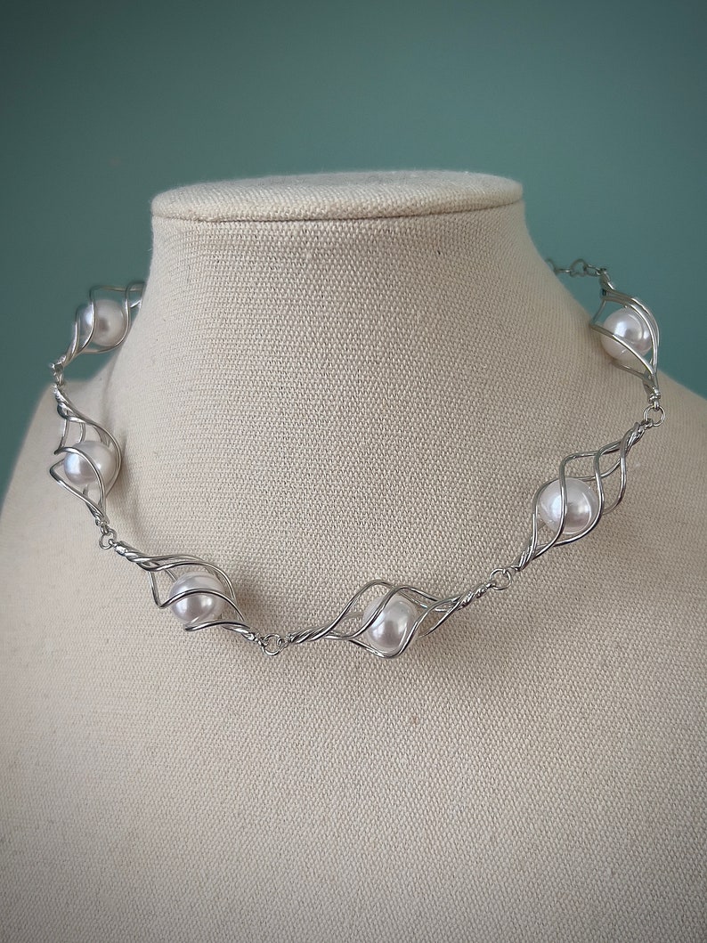 May include: A silver wire necklace with white pearls. The necklace is made of twisted wire that forms a delicate, organic shape. The pearls are evenly spaced and are held in place by the wire.