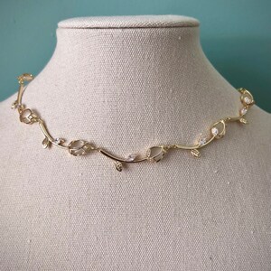 May include: A gold necklace with a delicate vine design and clear crystal accents. The necklace is displayed on a white fabric mannequin.