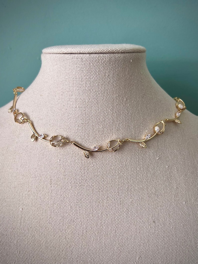 May include: A gold necklace with a delicate vine design and clear crystal accents. The necklace is displayed on a white fabric mannequin.