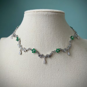 May include: A silver necklace with a delicate floral design featuring green and clear gemstones. The necklace has a pendant with a teardrop-shaped clear gemstone.
