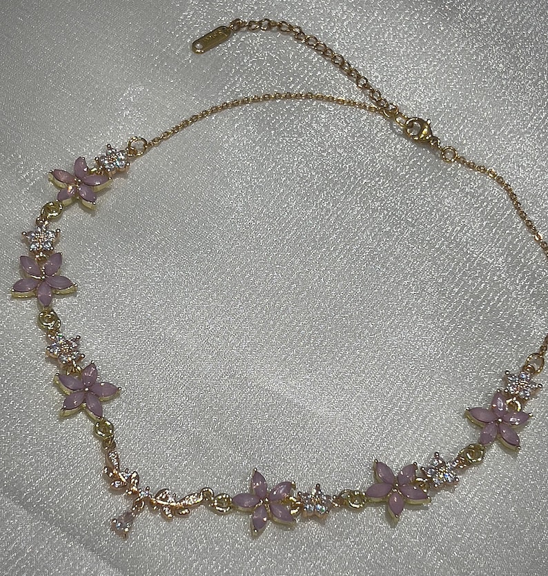May include: A gold chain necklace with pink flower accents and clear rhinestones. The necklace has a delicate design and is perfect for adding a touch of elegance to any outfit.