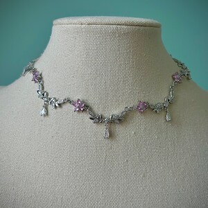 May include: A silver necklace with a delicate floral design and pink and clear crystals. The necklace features dangling crystal drops.