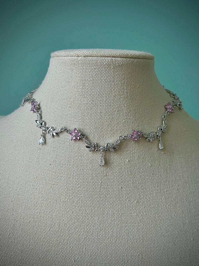 May include: A silver necklace with a delicate floral design and pink and clear crystals. The necklace features dangling crystal drops.
