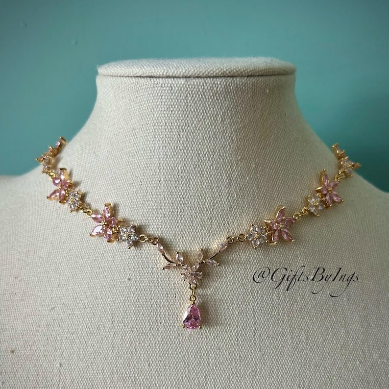 May include: A gold-tone necklace featuring a series of pink and clear crystal flower motifs connected by a delicate chain. The center of the necklace includes a dangling teardrop-shaped pink crystal pendant. The text @GiftsByIngs is visible.